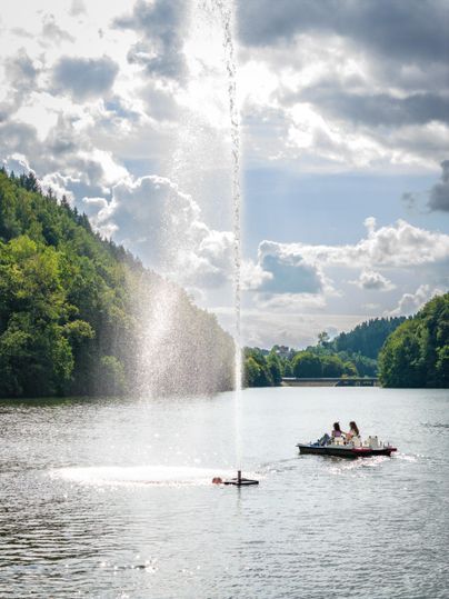 Un lac paisible avec une fontaine d'eau au milieu. À l'arrière-plan, des gens naviguent dans un petit bateau.