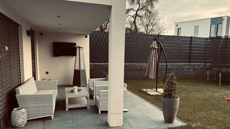 Une terrasse confortable avec un coin salon en rotin et une télévision à écran plat. En arrière-plan, un parasol est présent et le jardin est bien entretenu.