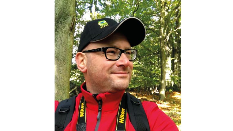 A man is wearing a red shirt and a baseball cap. He is standing in a forest with green trees in the background.