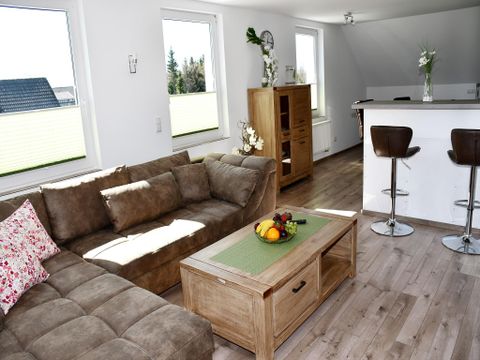 A bright living room with a beige sofa and a wooden table. In the background, there is a modern kitchenette.