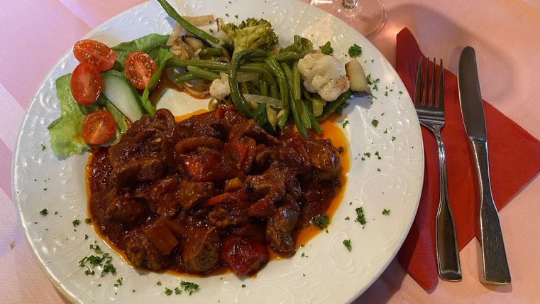 A plate with a hearty meat dish in sauce, accompanied by fresh vegetables and salad. Next to it are cutlery and a glass of wine.