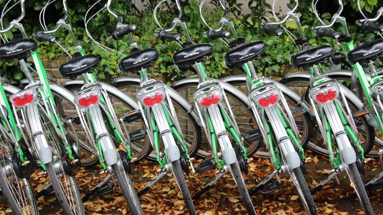 <p>In einer Reihe sind acht grüne Fahräder nebeneinander aufgereiht. Die nicht motorisierten Räder stehen auf ihren Fahrradständern, Lenker nach vorne, Gepäckträger hinten.</p>