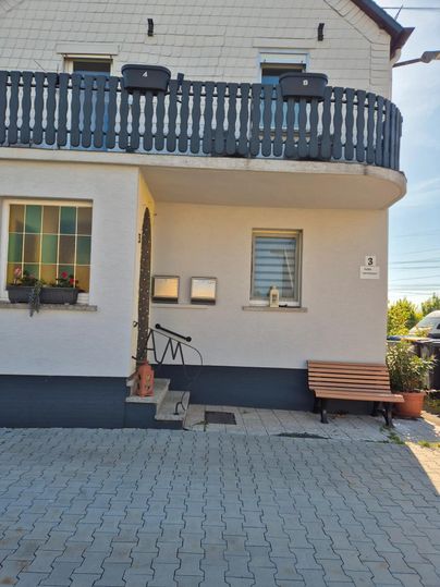 Ein Haus mit einem Balkon und einer Eingangstür. Vor dem Haus befindet sich eine Sitzbank und ein gepflasterter Bereich.