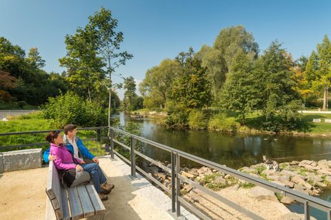 Deux personnes sont assises sur un banc au bord d'une eau calme. Entourées d'arbres et de verdure, elles profitent de la belle nature.