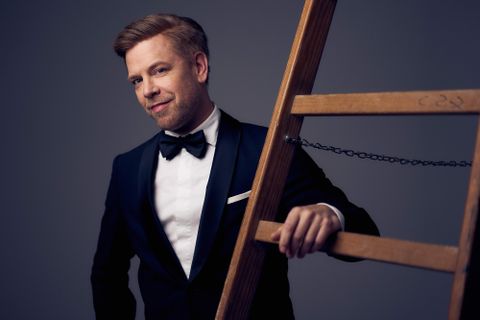 Eine Gruppe von sechs Musikern posiert in einem gemütlichen Raum. Sie stehen und sitzen um einen roten Sessel und tragen lässige Kleidung.