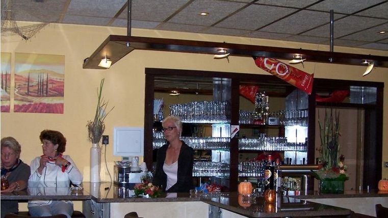 Een gezellige bar met een toog waar gasten zitten. Op de achtergrond zijn glazen en decoratieve planten zichtbaar.