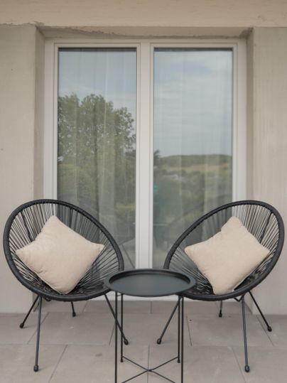 Een gezellig balkon met twee zwarte fauteuils en een tafeltje. De fauteuils zijn voorzien van lichte kussens en op de achtergrond zijn ramen met gordijnen te zien.