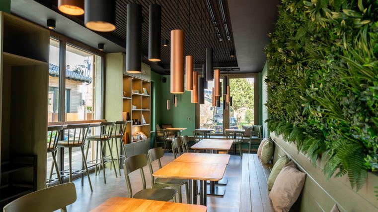 Een modern café met houten stoelen en tafels. De muren zijn versierd met planten, en er zijn grote ramen die veel licht binnenlaten.