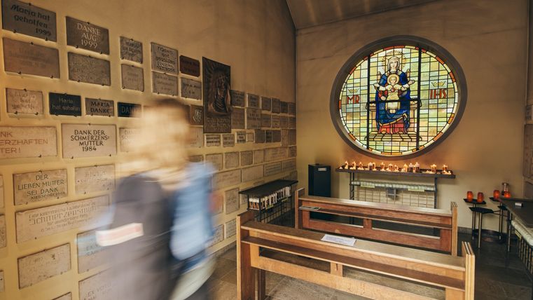 Small chapel with a person in motion in the foreground, surrounded by thanksgiving plaques on the walls. In the center, a round stained glass window with a figure of the Virgin Mary and an altar with burning candles