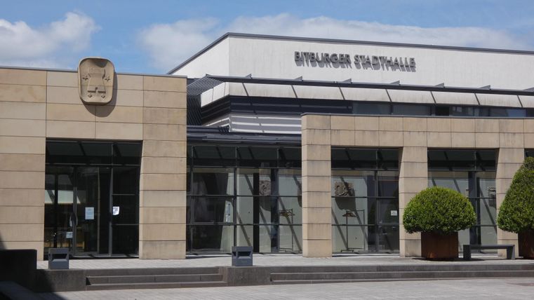 Ein modernes Gebäude mit Glasfassade und einem Eintragungszelt. Vor dem Gebäude stehen gepflegte Pflanzen.