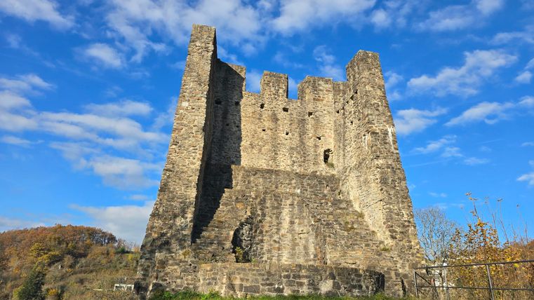 Eine alte Burgruine steht auf einem Hügel, umgeben von grünem Gras. Der Himmel ist blau mit wenigen Wolken.
