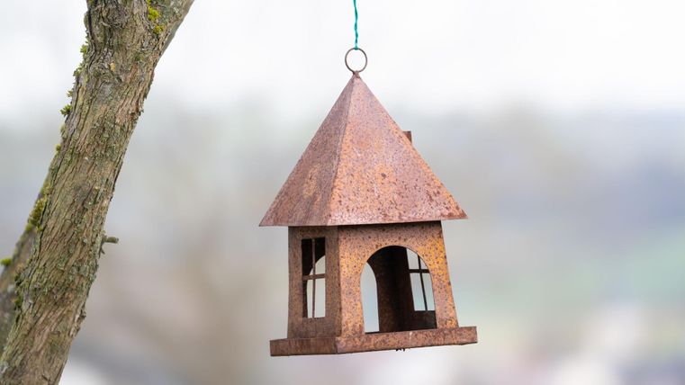 Ein rustikales Vogelhaus hängt an einem Baum. Es hat ein spitzes Dach und ist aus Metall gefertigt.