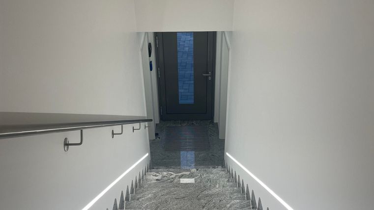 Eine moderne Treppe mit grauen Stufen und minimalistischem Design. Der Eingangsbereich ist hell beleuchtet und führt zu einer Tür.