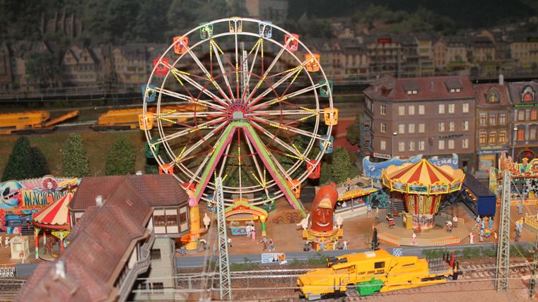 Ein kleines Jahrmarktmodell mit einem Riesenrad und einem Karussell. Im Hintergrund sind Gebäude und Züge zu sehen.