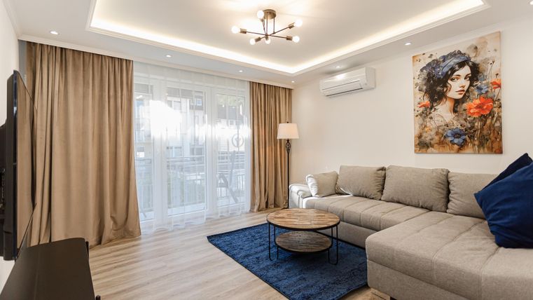 A modern living room with a gray sofa and a round table. A colorful artwork hangs on the wall, and large windows provide plenty of daylight.