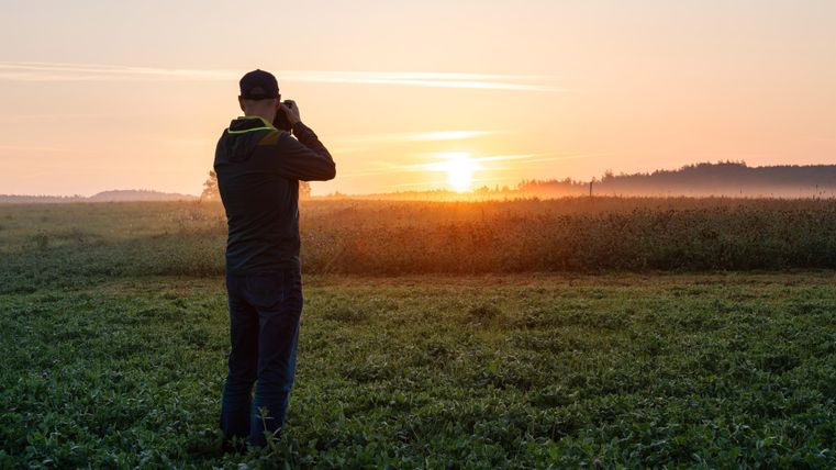 Ein Mann steht auf einem Feld und fotografiert einen Sonnenaufgang. Die Szene zeigt sanfte Farben und eine ruhige Morgenstimmung.
