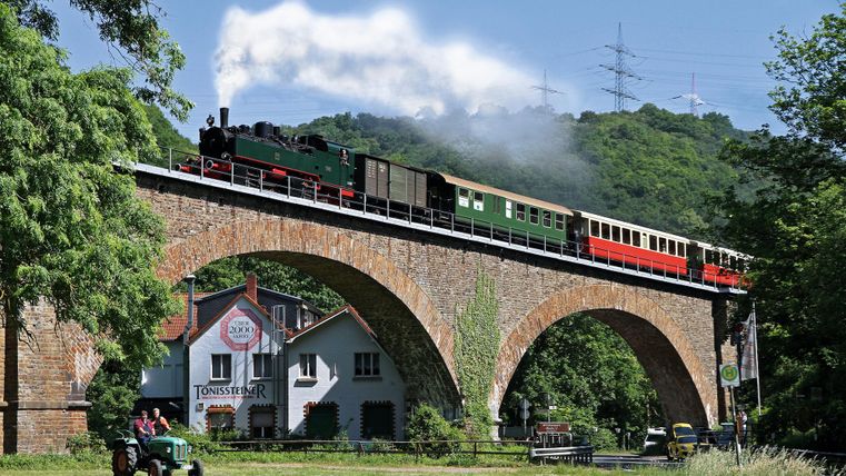Eine Dampflok fährt über eine große Steinbrücke. Im Hintergrund sind Bäume und ein Gebäude zu sehen.