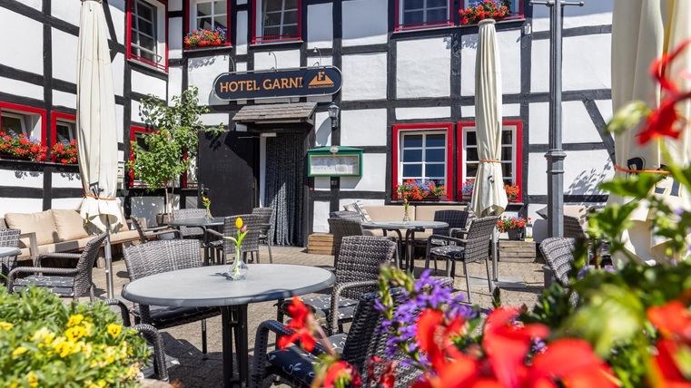 Een gezellige terrasje voor een hotel met mooie planten en bloemen. Er zijn tafels en stoelen die uitnodigen om te ontspannen.