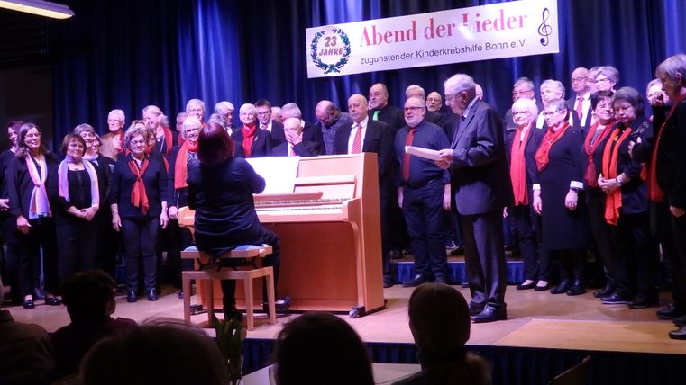 A choir is singing on a stage, accompanied by a piano. The singers are wearing red scarves and there is a festive atmosphere.