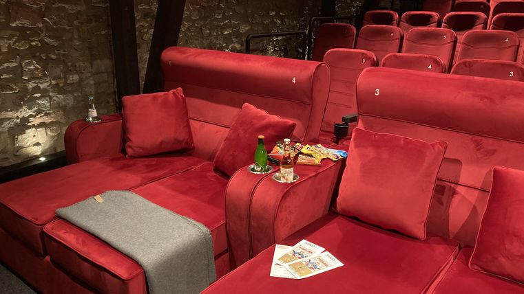 Een sfeervolle bioscoop met rode stoelen en stijlvolle bank-elementen. Snacks en drankjes zijn op tafel klaar.