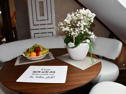 Une table élégante avec un bol plein de fruits frais et une plante en fleurs. Sur la surface de la table, il y a une plaque avec l'inscription : "Manger ne fait pas grossir, manger ne compte que le bon talent !"