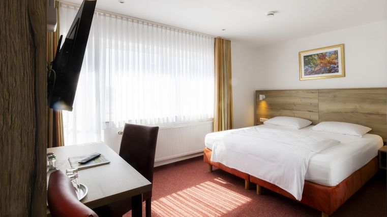 A bright hotel room with a large double bed and a desk. The room has large windows that let in a lot of daylight.