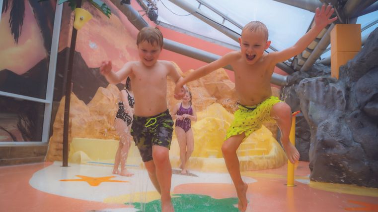 Deux enfants joyeux sautent dans un parc aquatique. En arrière-plan, on peut voir des équipements de jeu colorés et une atmosphère de vacances détendue.