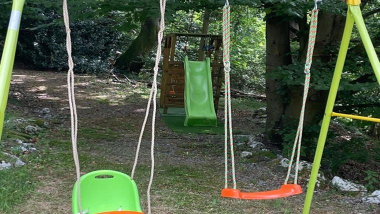 A swing with two seats is located in a green, wooded area. In the background, a slide can be seen.