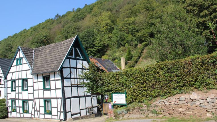 Ein weißes Fachwerkhaus mit schwarzem Dach steht an einer Straße. Im Hintergrund sind grüne Hügel und Bäume zu sehen.