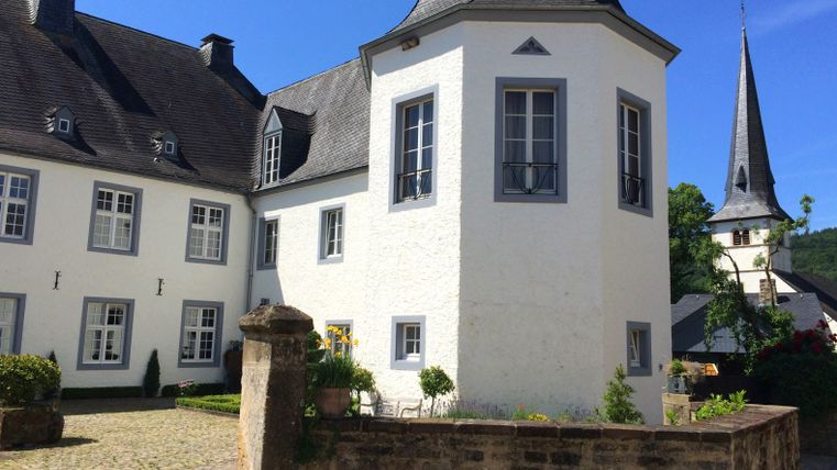 White castle building with gray roof and tower, surrounded by garden and stone wall, under a blue sky.