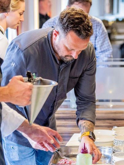 Een kok werkt geconcentreerd aan een gerecht, terwijl een collega hem bijstaat. Op de achtergrond zijn andere mensen en keukengerei te zien.