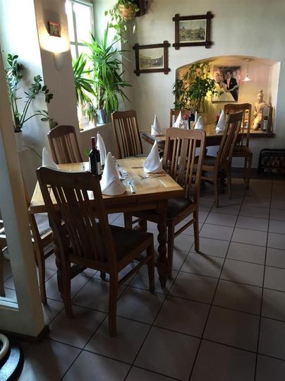 A cozy restaurant with wooden tables and chairs, white napkins, plants, and wall paintings. An inviting, bright room.