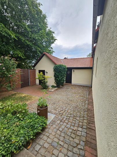 Een rustige binnenplaats met een geplaveide oppervlakte en kleurrijke planten. Op de achtergrond zijn een klein huis en bomen te zien.