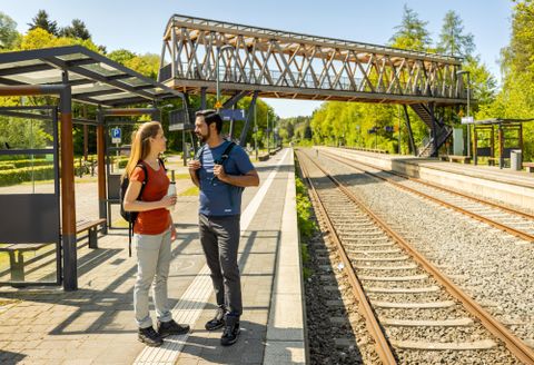 A train station outdoors with two travelers talking to each other. In the background, tracks and a pedestrian crossing can be seen.