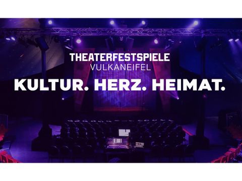 Ein Veranstaltungssaal mit roten Stühlen und einer Bühne im Hintergrund. Die Beleuchtung ist in Violett gehalten und vermittelt eine einladende Atmosphäre.