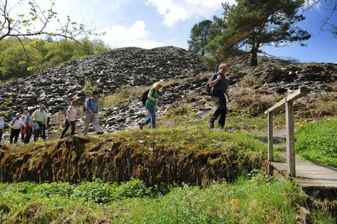 Eine Gruppe von Wanderer ist auf einem schmalen Pfad unterwegs. Im Hintergrund ist ein steiler, dunkler Schieferberg mit vereinzelter Vegetation zu sehen. 