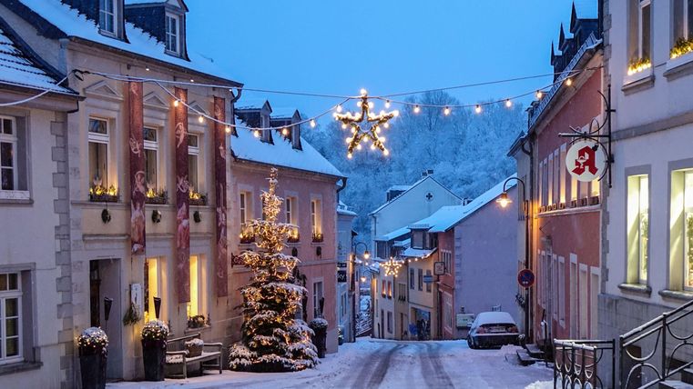 Une rue enneigée avec des maisons décorées de manière festive. Des lumières et un sapin de Noël créent une atmosphère chaleureuse au crépuscule.