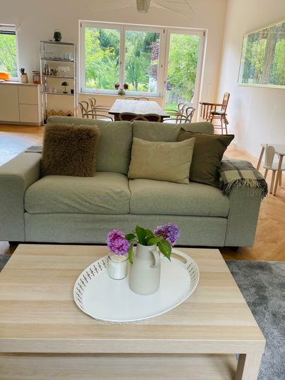 Un salon confortable avec un canapé gris et une table basse en bois. Sur la table se trouve un vase avec des fleurs fraîches et les fenêtres offrent une vue sur la verdure.