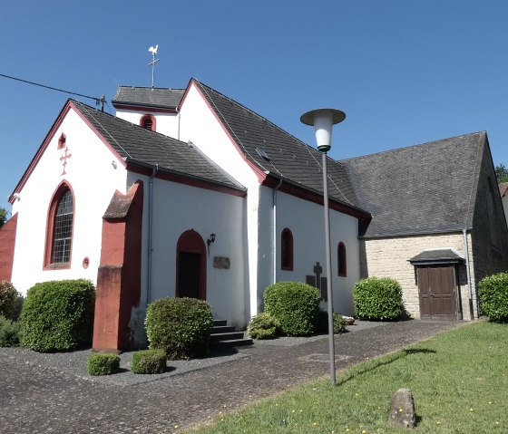 Weiße Kirche mit roten Akzenten und grauem Dach in Dahlem, umgeben von gepflegten Büschen und Bäumen unter klarem, blauem Himmel., © start#99!