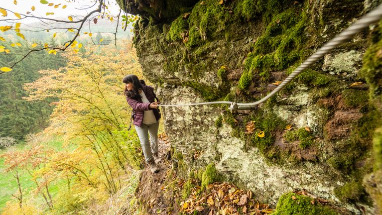 Een persoon loopt over een rotsachtig, met mos bedekt pad, terwijl hij zich vasthoudt aan een touw. Herfstbomen op de achtergrond.