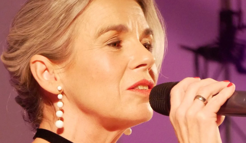 Une femme aux cheveux gris chante dans un microphone. Elle porte des vêtements élégants et des boucles d'oreilles.