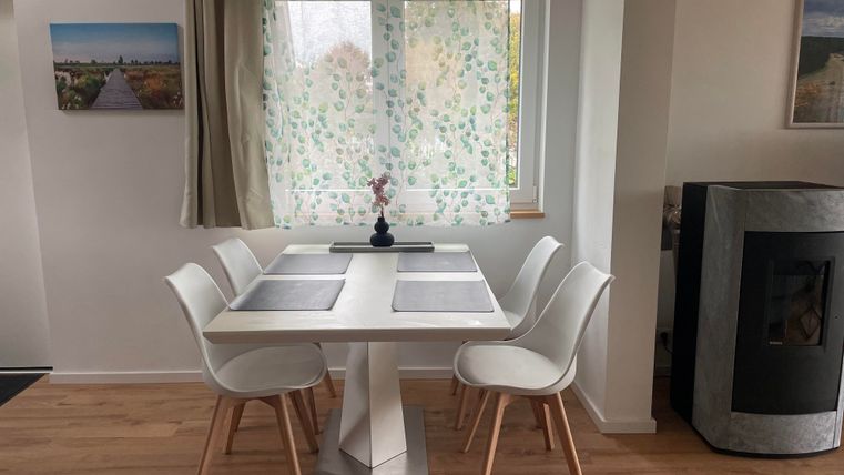 Ein heller Essbereich mit einem weißen Tisch und fünf Stühlen. Vor einem Fenster mit einem dekorativen Vorhang steht eine kleine Vase mit Blumen.