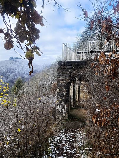 Une ancienne structure en pierre dans un paysage hivernal, entourée d'arbres et de neige. À l'arrière-plan, des collines douces se dessinent sous un ciel nuageux.