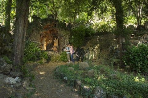 Een pittoreske tuin met weelderig groen en oude stenen. Twee personen staan voor een decoratieve niche te midden van de natuur.