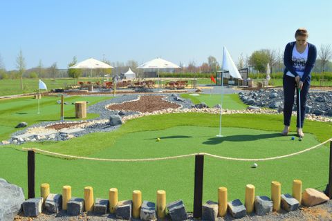 A woman is playing mini-golf on a beautifully designed course with green grass and stones. In the background, there are some pavilions and trees visible.