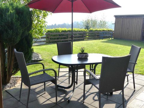 Een uitnodigende buitenruimte met een ronde tafel en vier stoelen onder een rode parasol. De tuin is groen en goed onderhouden, ideaal voor ontspannen uurtjes buiten.