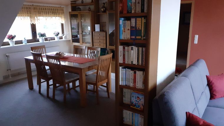 A cozy living space with a dining table and chairs. On the right, there is a shelf full of books and a gray couch.