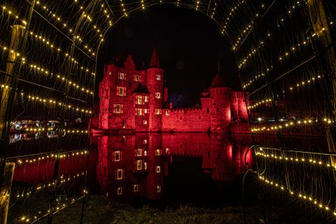 Eine beeindruckende Burg, die nachts rot beleuchtet ist. Sie spiegelt sich im Wasser und ist von funkelnden Lichtern umgeben.