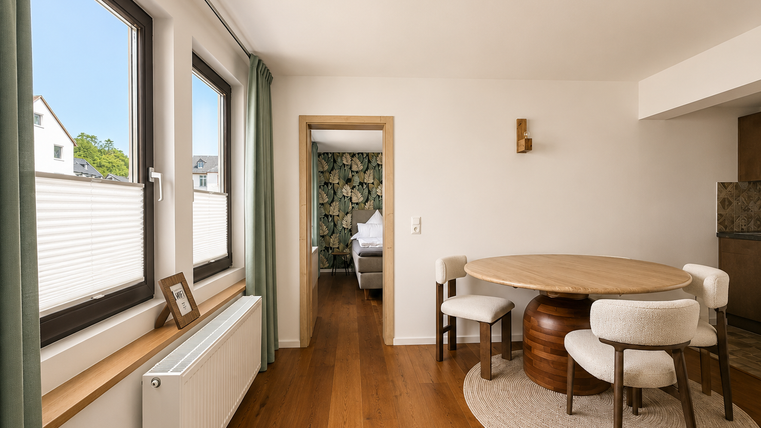 Een moderne woonkamer met een ronde tafel en elegante stoelen. Door grote ramen valt er veel daglicht naar binnen en creëert het een lichte sfeer.