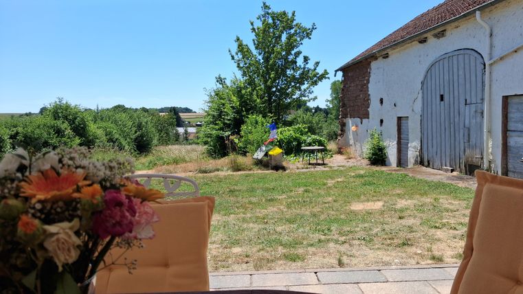Une prairie verte avec un arbre ombragé et un ancien bâtiment en arrière-plan. Au premier plan, il y a une table avec des fleurs.
